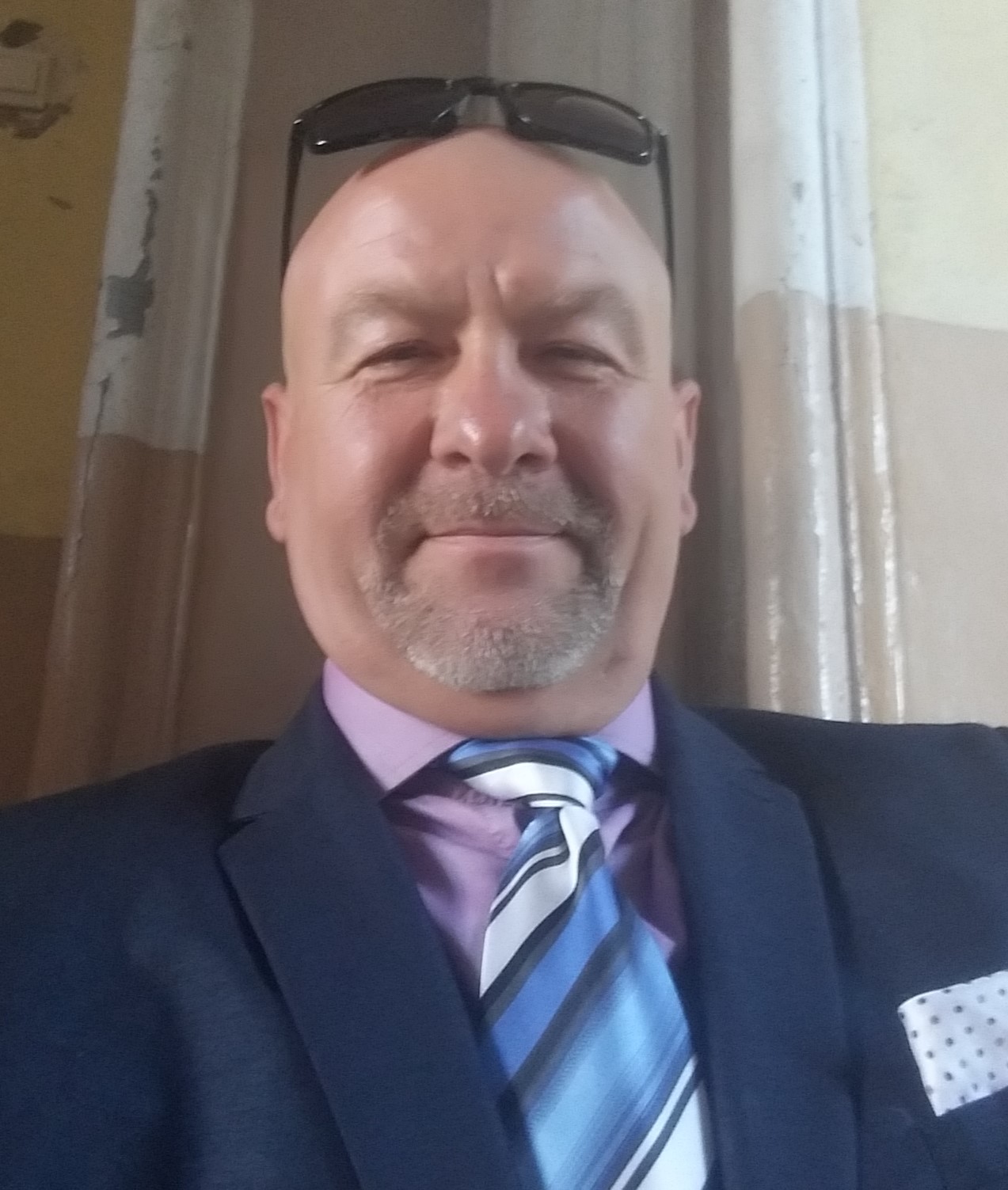 Portret uśmiechniętego mężczyzny w garniturze, z okularami na głowie, na tle kolumn w odcieniach beżu i brązu.