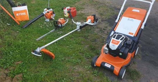 Zestaw sprzętu ogrodniczego marki Stihl na trawie: kosiarka, dmuchawa, pilarka, nożyce do żywopłotu i podkaszarka.