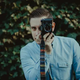 Młody mężczyzna z aparatem Canon zasłaniającym część twarzy, w tle zielone liście, na palcu złoty pierścionek, koszula jeansowa.