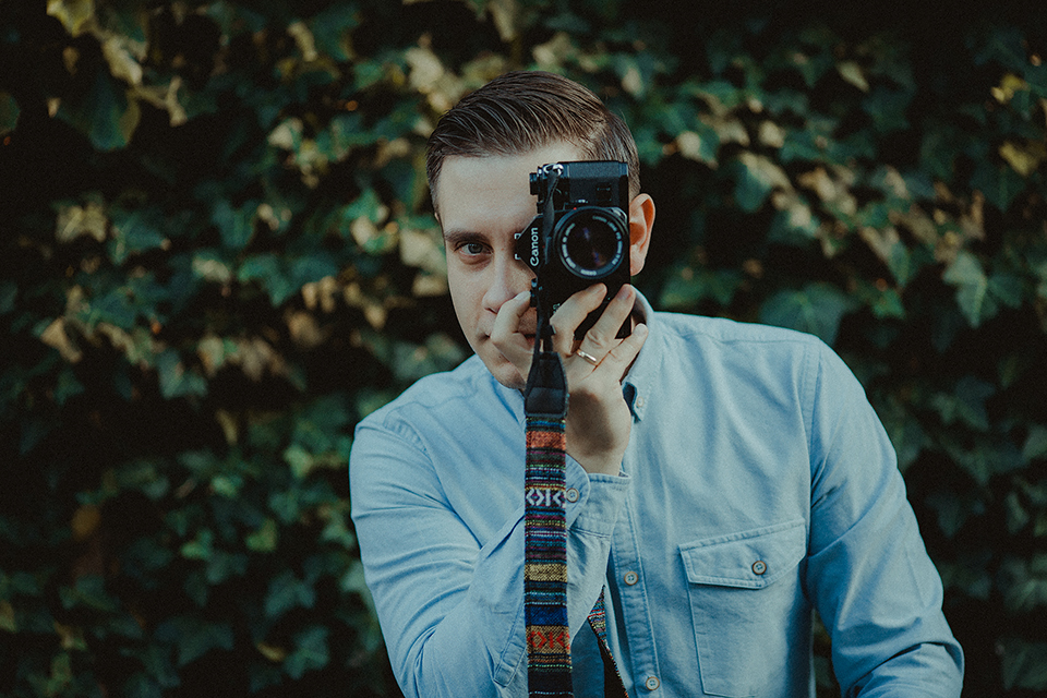 Młody mężczyzna z aparatem Canon zasłaniającym część twarzy, w tle zielone liście, na palcu złoty pierścionek, koszula jeansowa.