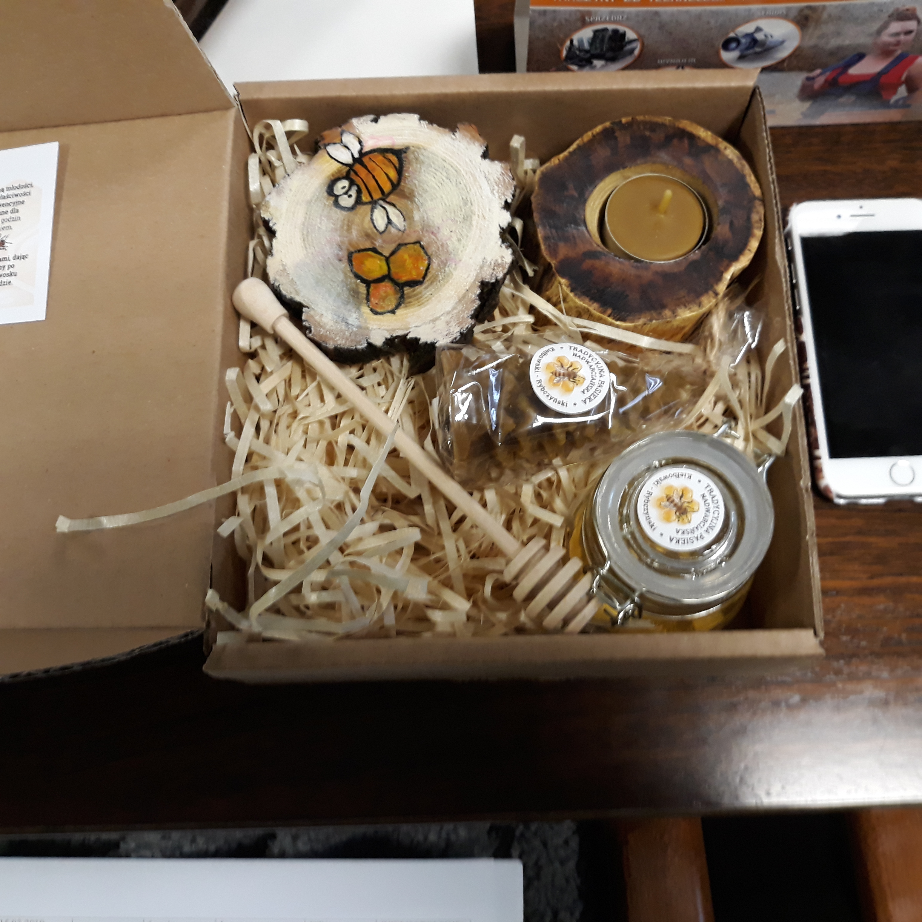 Otwarty kartonowy kosz prezentowy z produktami pszczelarskimi: słoik miodu z drewnianym nabierakiem, świeczka w drewnianej oprawie, kostka wosku pszczelego oraz plaster drewna z namalowaną pszczołą...
