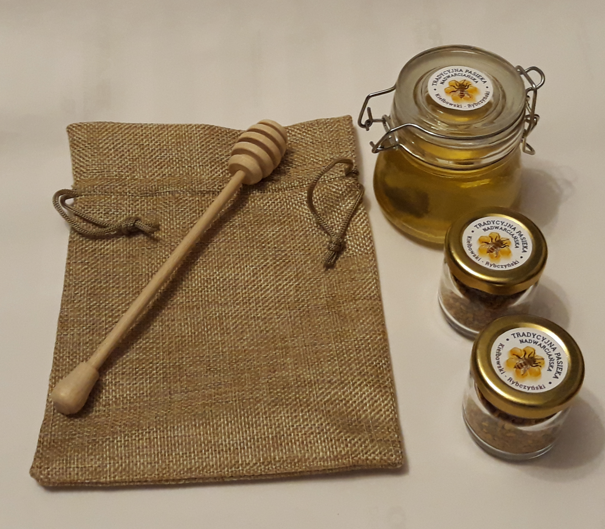 Zestaw prezentowy z miodem i pyłkiem pszczelim w szklanych słoiczkach, zapakowany w jutowy woreczek z drewnianym nabierakiem do miodu, etykiety z logo 'Tradycyjna Pasieka Nadwarciańska'.
