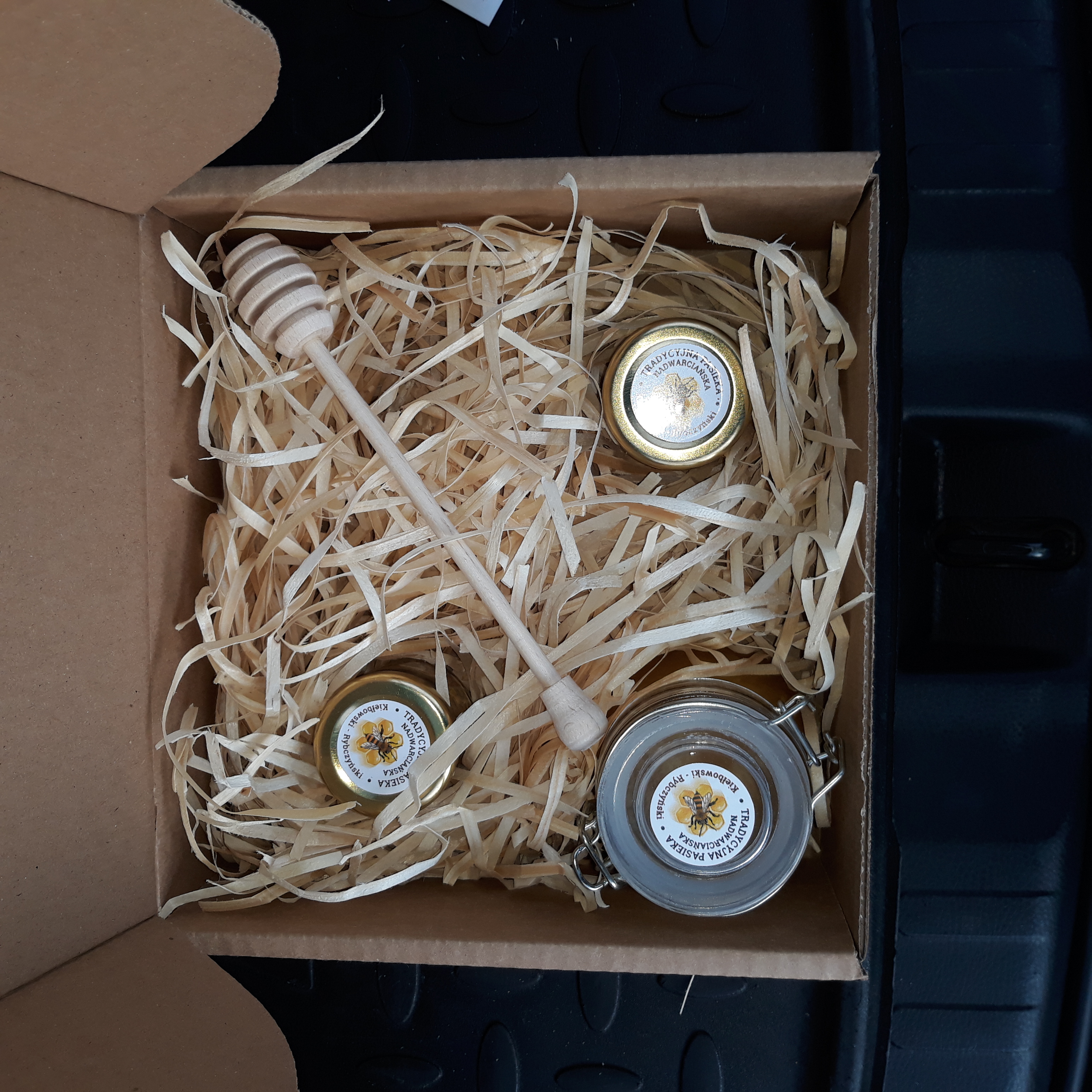 Otwarty kartonowy prezentowy z trzema słoiczkami miodu z etykietami 'Tradycyjna Pasieka Nadwarciańska' i drewnianym nabierakiem na wiórkach papierowych.