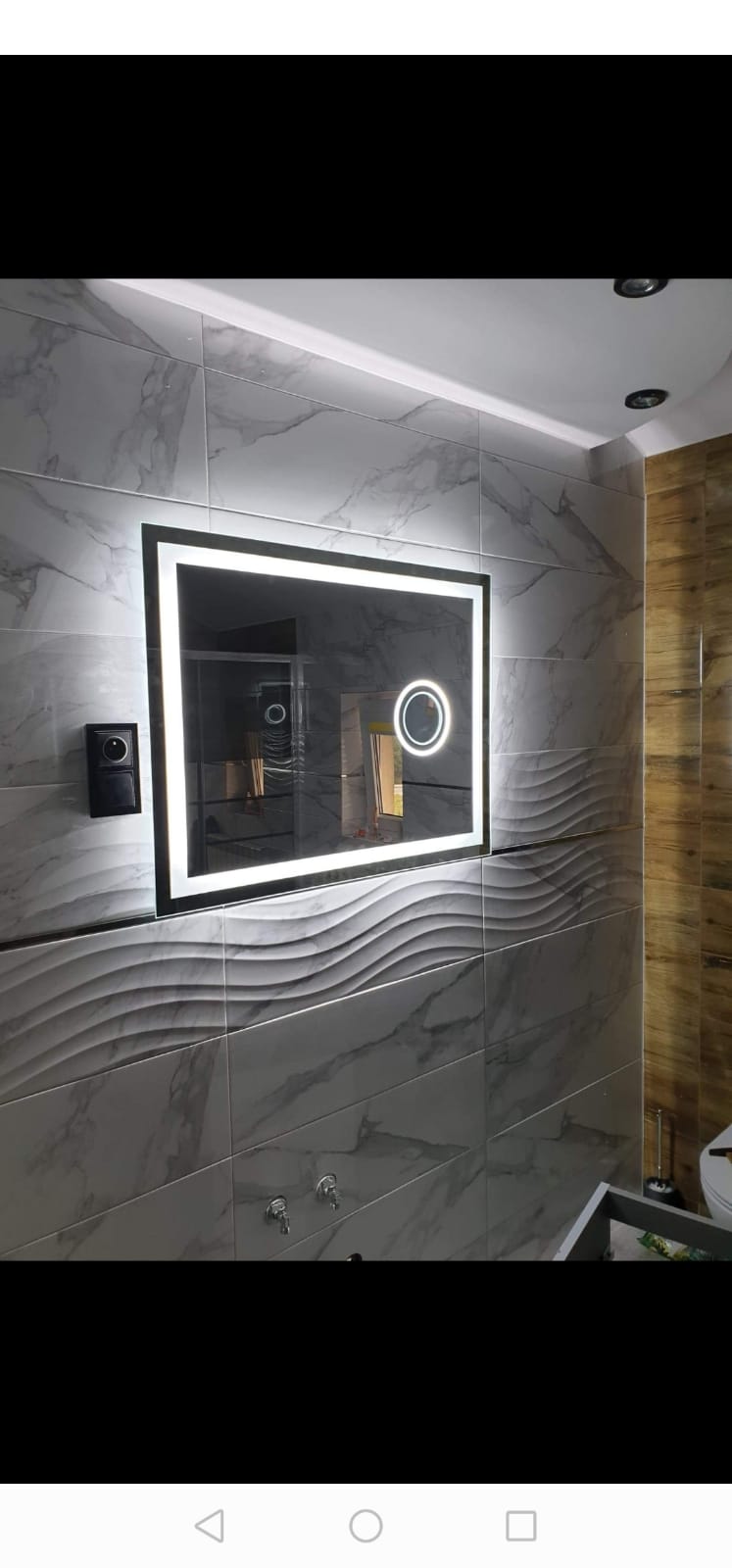 Nowoczesna łazienka z podświetlanym lustrem w ramie, marmurowe płytki z falistym wzorem, drewniane panele po prawej stronie i czarny włącznik światła.