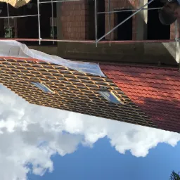 Częściowo pokryty dach z czerwonej dachówki z dwoma oknami dachowymi, widoczna konstrukcja drewniana i folia ochronna, w tle fragment murowanej ściany budynku na rusztowaniu.