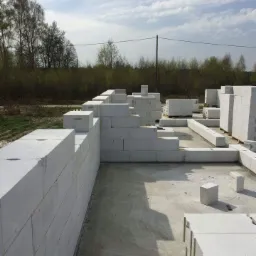 Wznoszenie ścian fundamentowych z bloczków betonu komórkowego na wylewce, widok z poziomu gruntu, w tle drzewa bezlistne.