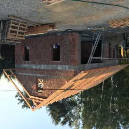 Wznoszenie dwukondygnacyjnego budynku z czerwonej cegły z widoczną konstrukcją dachu z drewnianych belek, palety z materiałami budowlanymi i betoniarka w tle.