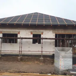 Dom w trakcie budowy: murowane ściany z białych bloczków, częściowo ocieplony dach z widocznymi łatami, rusztowania wokół budynku.