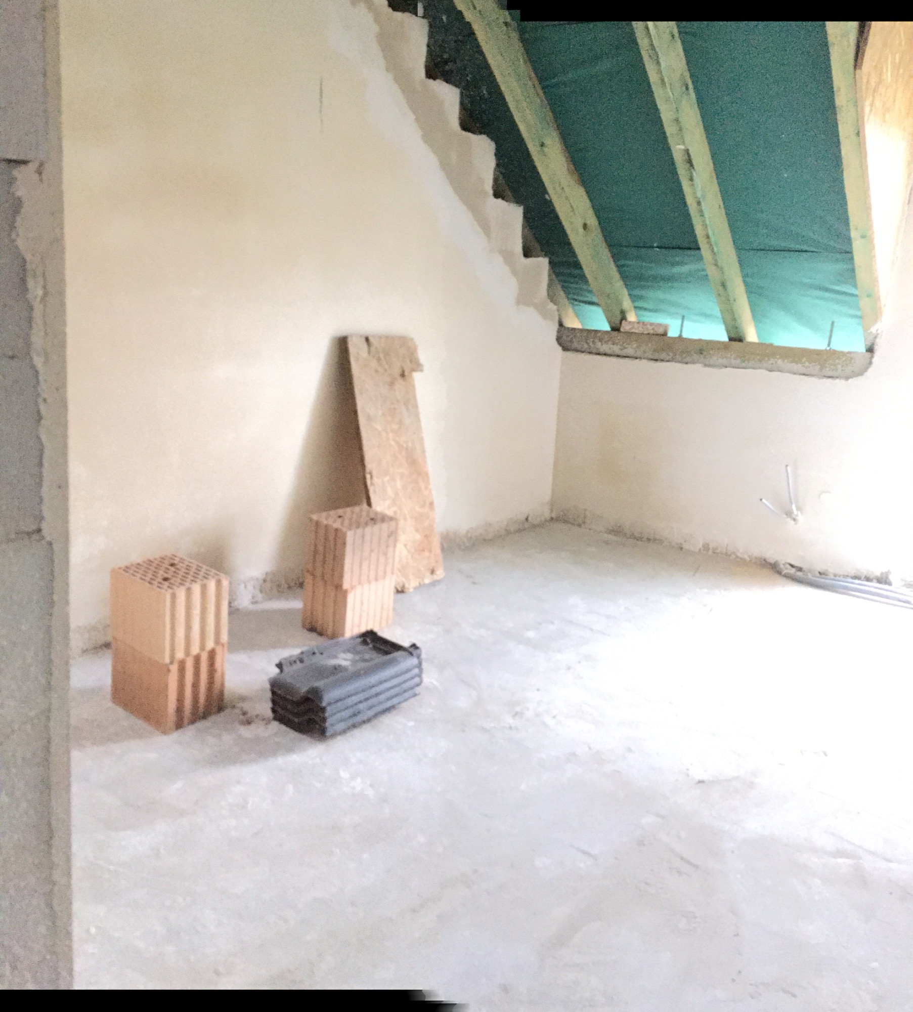 Surowy stan poddasza z oknem dachowym w trakcie budowy, widoczne pustaki, deska i plastikowe pojemniki na podłodze.