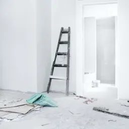 Pokój w trakcie remontu: białe ściany, drabina oparta o ścianę, resztki karton-gipsu i narzędzia na podłodze, otwarte drzwi do kolejnego pomieszczenia.
