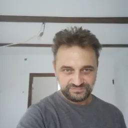 Mężczyzna z brodą w szarej koszulce na tle białej ściany z drewnianymi belkami sufitowymi i framugą drzwi w trakcie remontu.