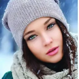 Portret kobiety w zimowej scenerii, w szarej czapce i szaliku, z delikatnym makijażem i zielonymi oczami, na tle padającego śniegu i rozmytego krajobrazu.