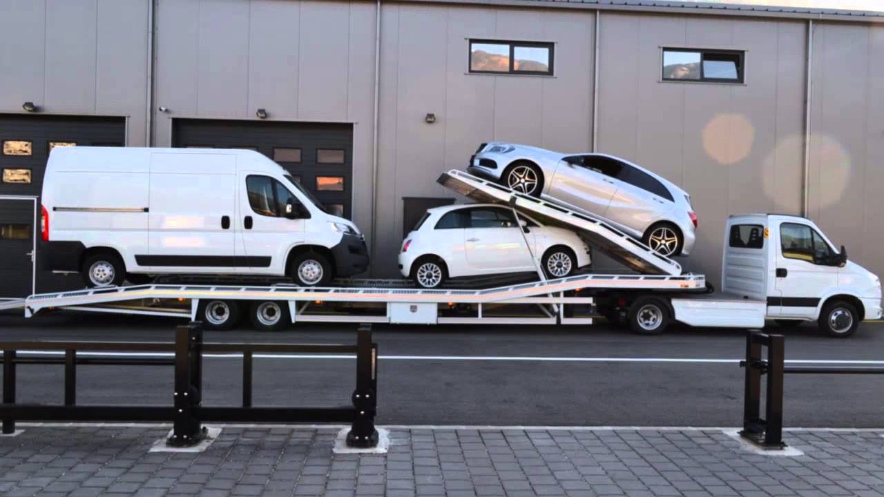 Laweta z trzema białymi pojazdami: furgonetką, Fiatem 500 i Mercedesem klasy A, na tle szarego budynku z bramami garażowymi.