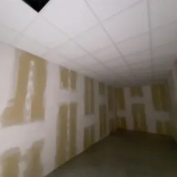 Pomieszczenie w trakcie prac przygotowawczych do malowania, widoczne fragmenty ścian z nałożoną warstwą szpachli, sufit kasetonowy.