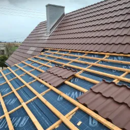 Montaż brązowej blachodachówki na drewnianej konstrukcji z folią hydroizolacyjną, widoczny komin i fragment zabudowy w tle.