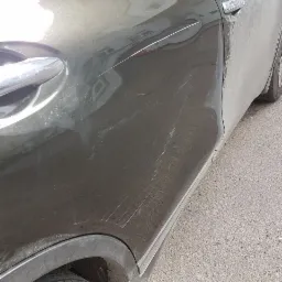 Błotnisty, oliwkowozielony samochód z licznymi zarysowaniami na drzwiach i błotniku, widoczne ślady po kontakcie z gałęziami lub innymi pojazdami.