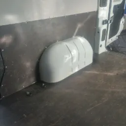 Wnętrze furgonetki z zamontowaną płytą ochronną na ścianach i nadkolem. Widoczne mocowania w podłodze i narzędzie do obkurczania folii w tle.