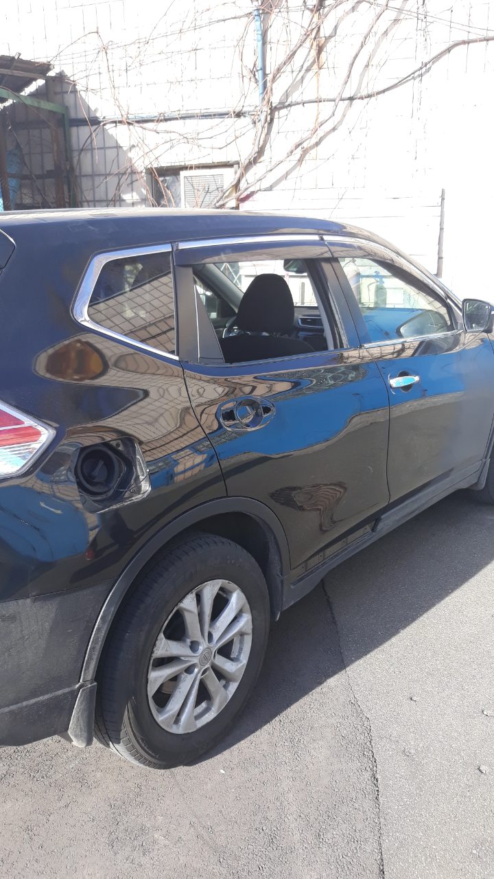 Częściowy widok boku czarnego samochodu, z otwartą klapką wlewu paliwa, zaparkowanego na szarym asfalcie, z budynkiem w tle.