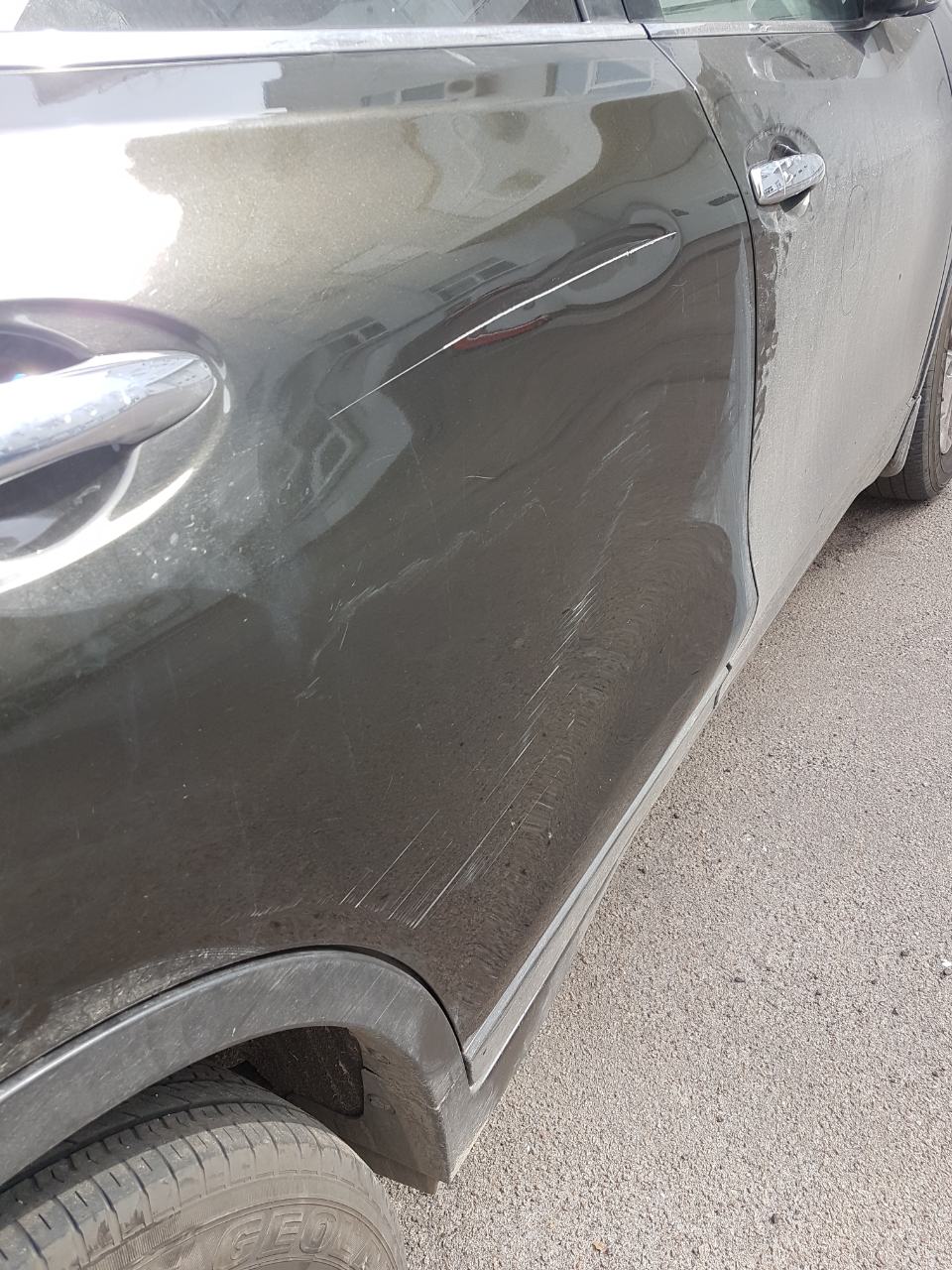 Błotnisty, oliwkowozielony samochód z licznymi zarysowaniami na drzwiach i błotniku, widoczne ślady po kontakcie z gałęziami lub innymi pojazdami.