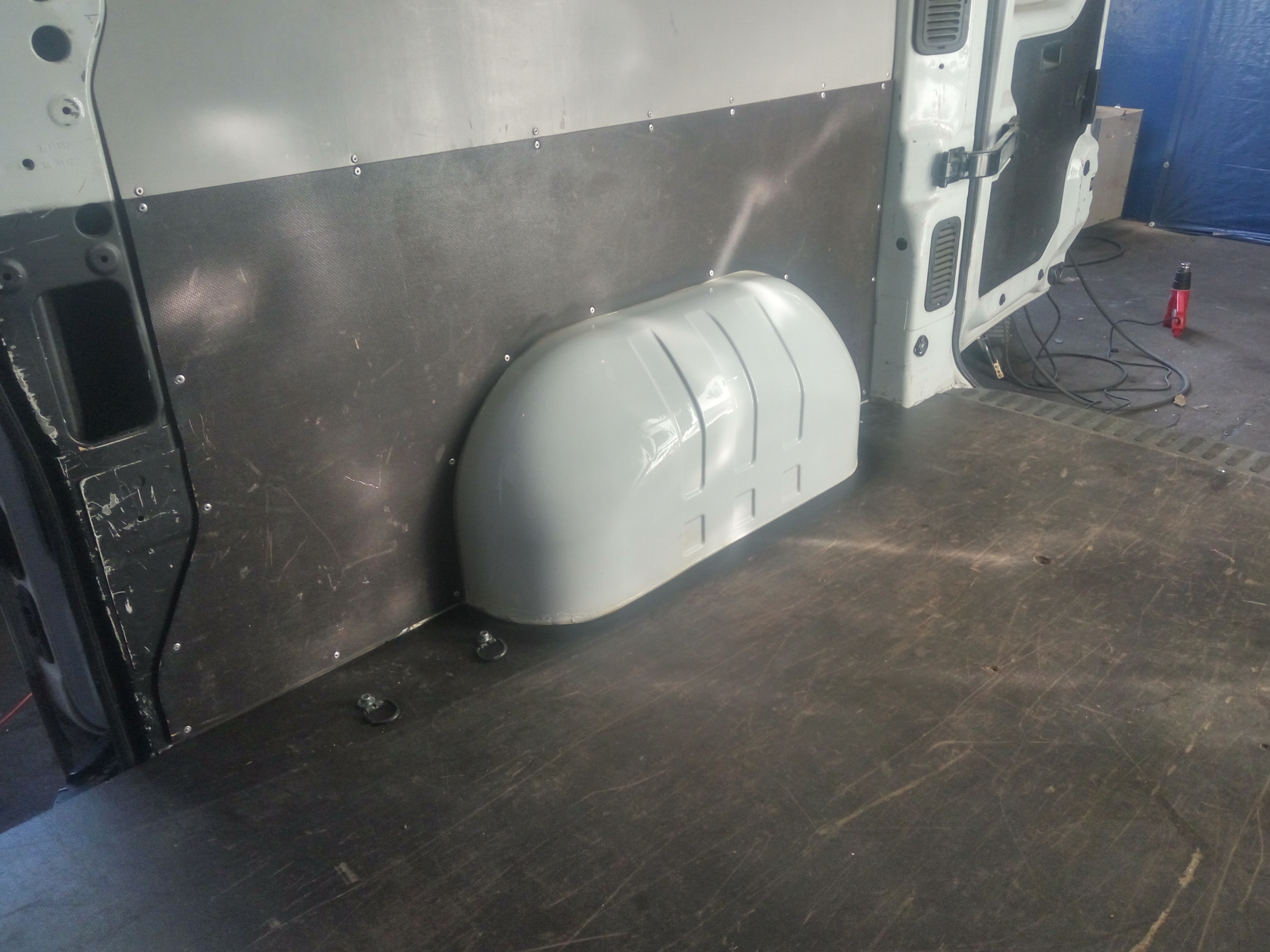 Wnętrze furgonetki z zamontowaną płytą ochronną na ścianach i nadkolem. Widoczne mocowania w podłodze i narzędzie do obkurczania folii w tle.