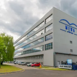 Szary budynek biurowy z dużą ilością okien i niebieskim logo 'MEWA' na fasadzie, otoczony zielenią i zaparkowanymi samochodami.