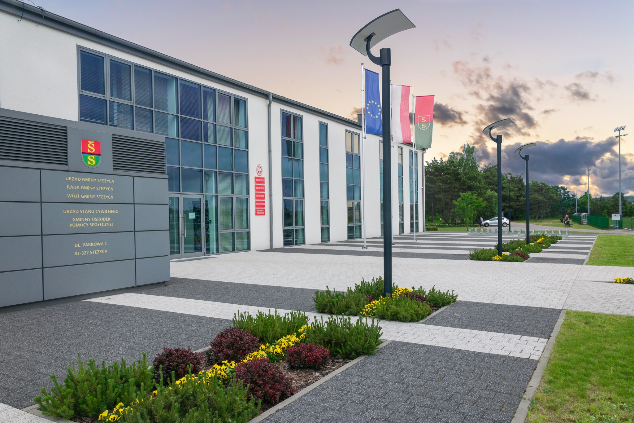 Budynek Urzędu Gminy Steżyca z flagami Polski, Unii Europejskiej i gminy, widoczny z zewnątrz, z uporządkowanym otoczeniem i elementami małej architektury.