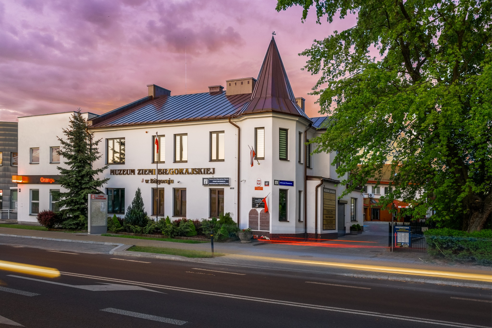 Muzeum Ziemi Biłgorajskiej o zmierzchu, biały budynek z wieżyczką, polskie flagi, ślady świateł samochodów na ulicy, fioletowe niebo.