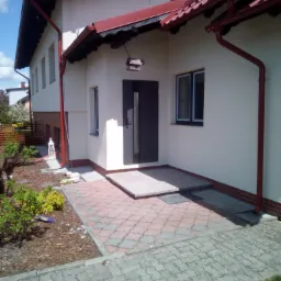 Elewacja domu jednorodzinnego z wejściem, widoczne czerwone rynny i dachówka, jasna elewacja, kostka brukowa i fragment ogrodu z ozdobnymi krzewami.