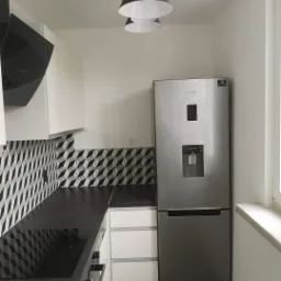 Nowoczesna kuchnia z czarnym blatem, geometrycznymi płytkami na ścianie, srebrną lod&oacute;wką Samsung i designerskimi lampami wiszącymi w kształcie stożk&oacute;w.