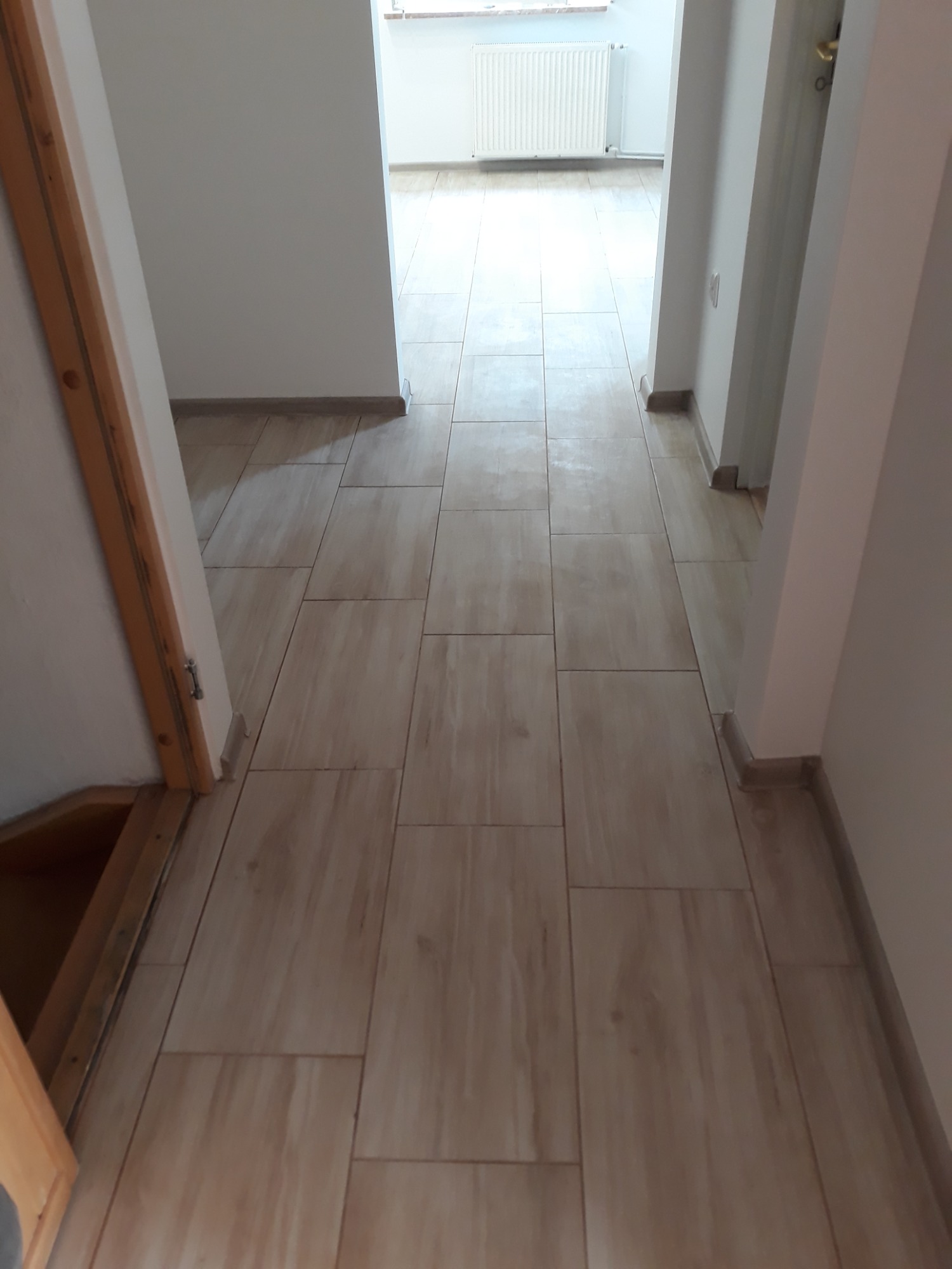 Wyłożona płytkami podłoga w korytarzu z widocznym fragmentem drewnianej ościeżnicy drzwi po lewej stronie i grzejnikiem w oddali.
