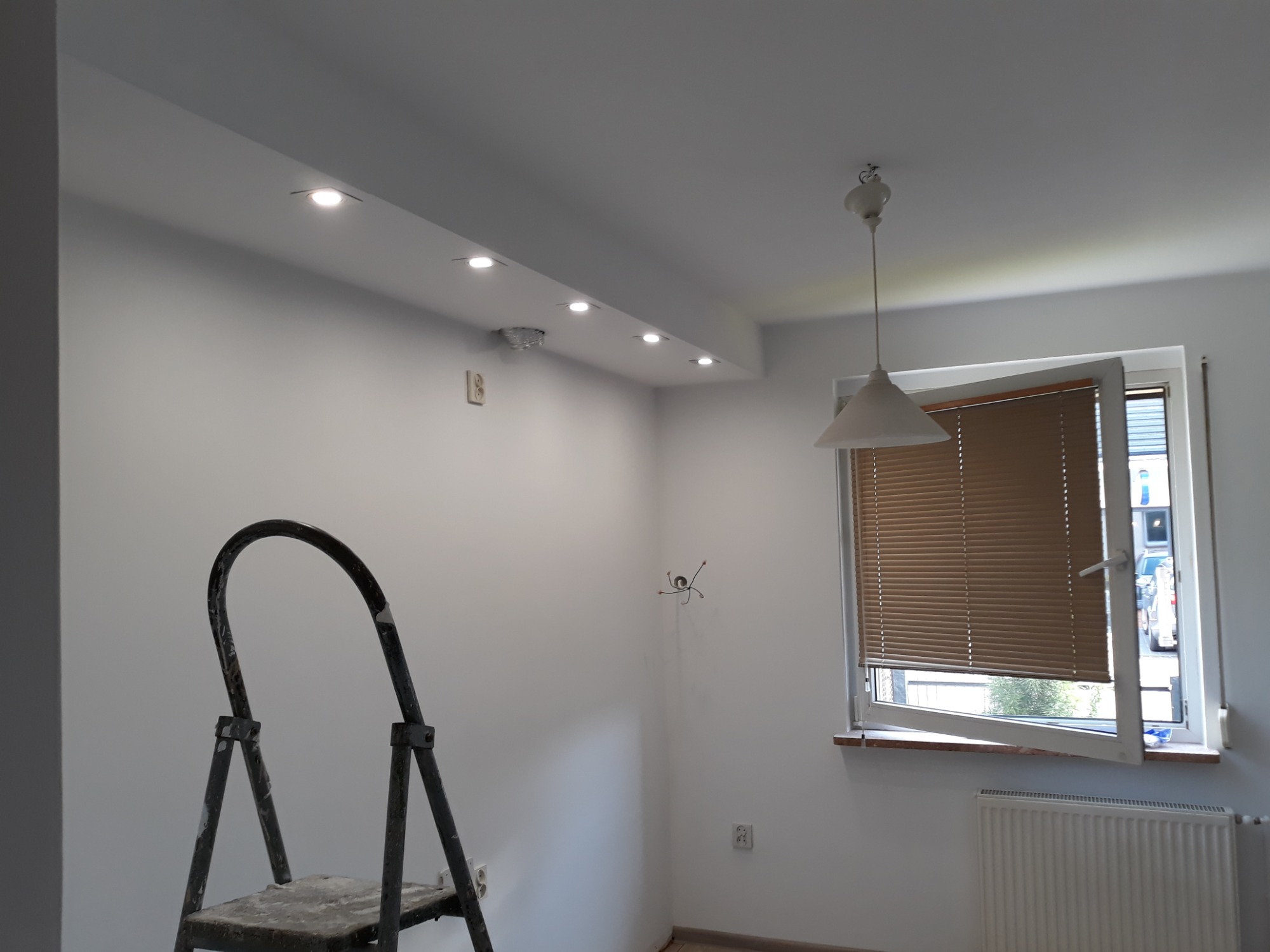 Pomieszczenie po malowaniu na biało, widoczna drabina malarska, zamontowane oświetlenie punktowe w suficie podwieszanym oraz okno z żaluzjami.