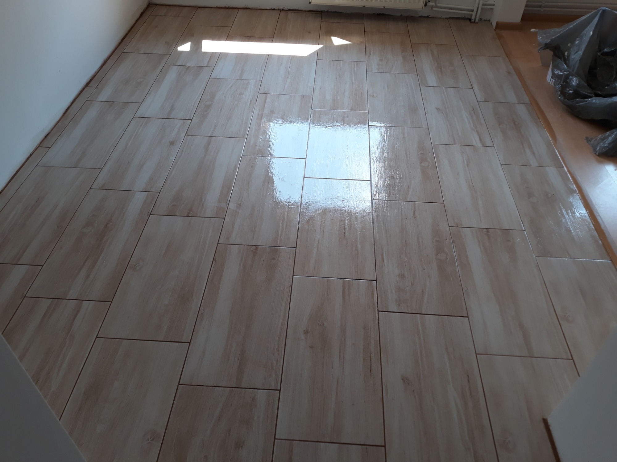 Ułożone jasnobeżowe płytki podłogowe imitujące drewno, ułożone pod kątem, z widocznym odbiciem światła słonecznego w centralnej części pomieszczenia.