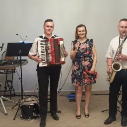 Zespół muzyczny w składzie: akordeonista, wokalistka z tamburynem i saksofonista, stojący przed sprzętem nagłośnieniowym i oświetleniowym.