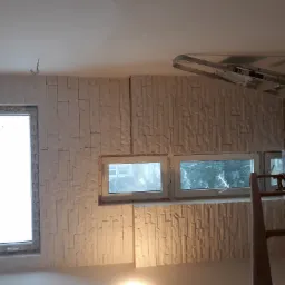 Ściana wykończona panelami imitującymi kamień, z dwoma oknami o różnej wielkości, widoczna drabina malarska i wystający kabel ze ściany.