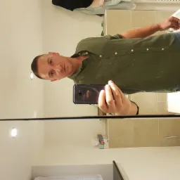 Mężczyzna robi selfie w lustrze łazienki z beżowymi płytkami. W tle umywalka i dozownik mydła. Widoczne odbicie telefonu w lustrze.