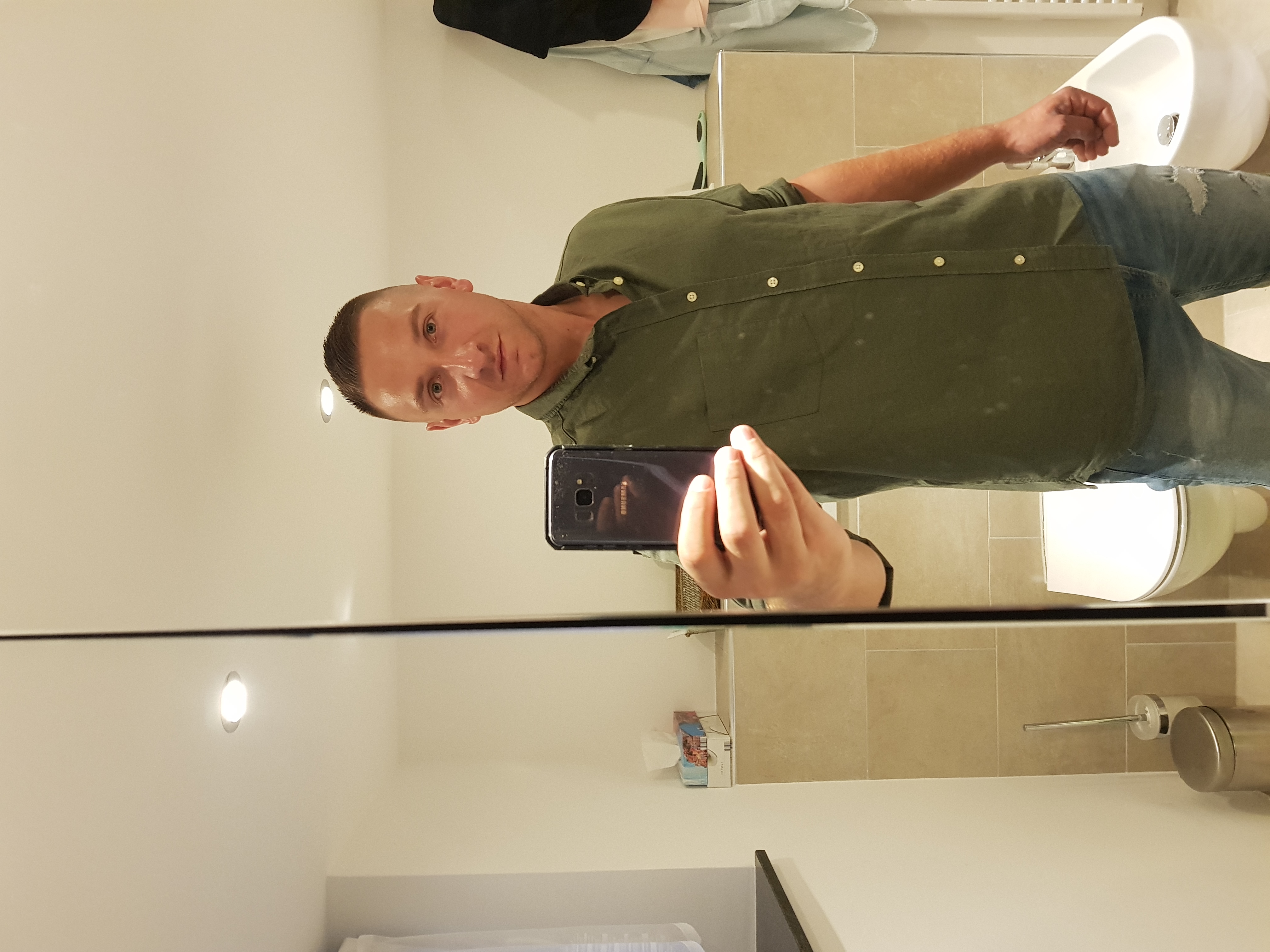 Mężczyzna robi selfie w lustrze łazienki z beżowymi płytkami. W tle umywalka i dozownik mydła. Widoczne odbicie telefonu w lustrze.