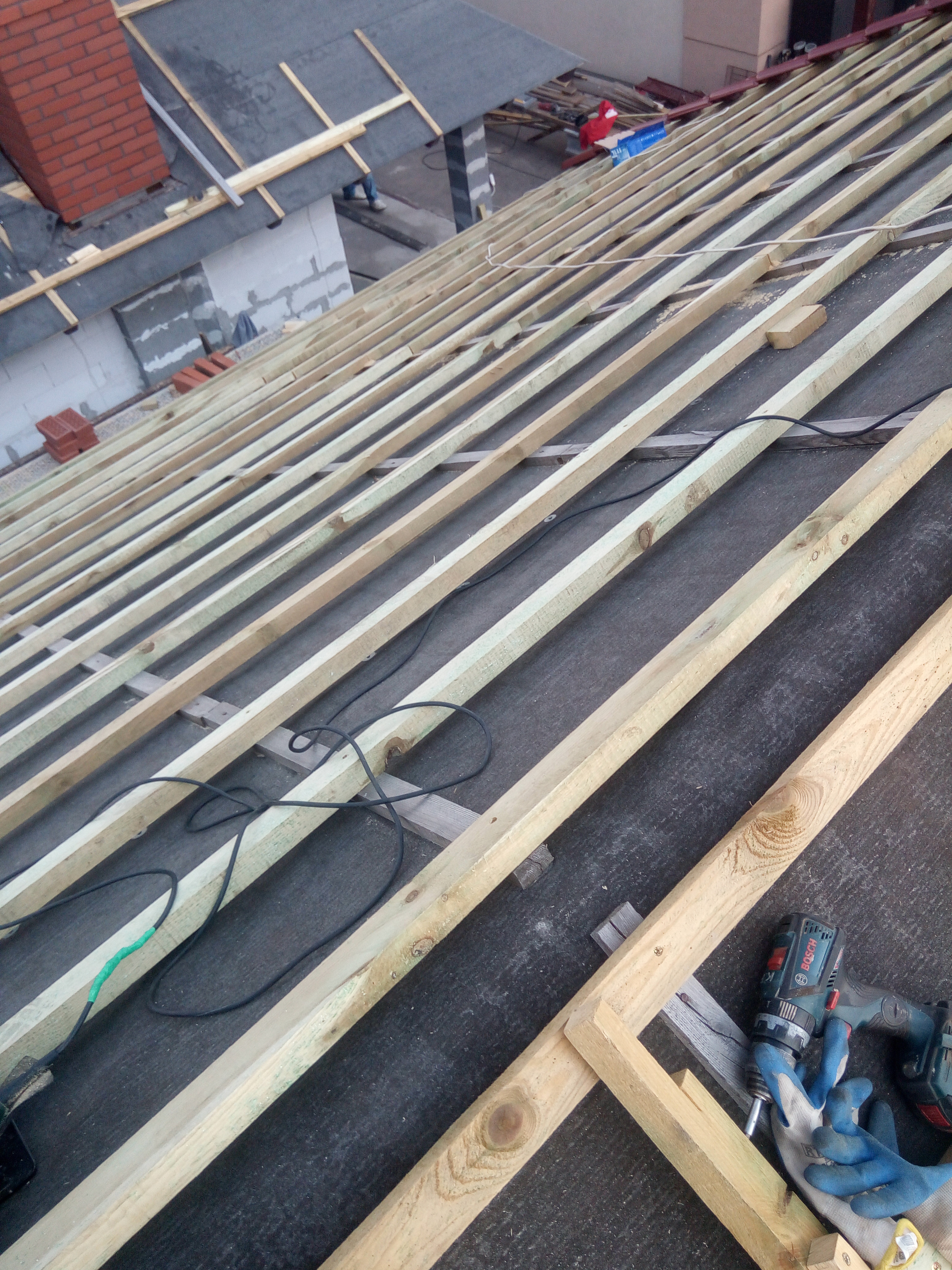 W trakcie budowy więźby dachowej w Nowym Mieście Lubawskim, widoczne drewniane elementy konstrukcyjne, czarna membrana dachowa, narzędzia pracy: wkrętarka Bosch i rękawice robocze.