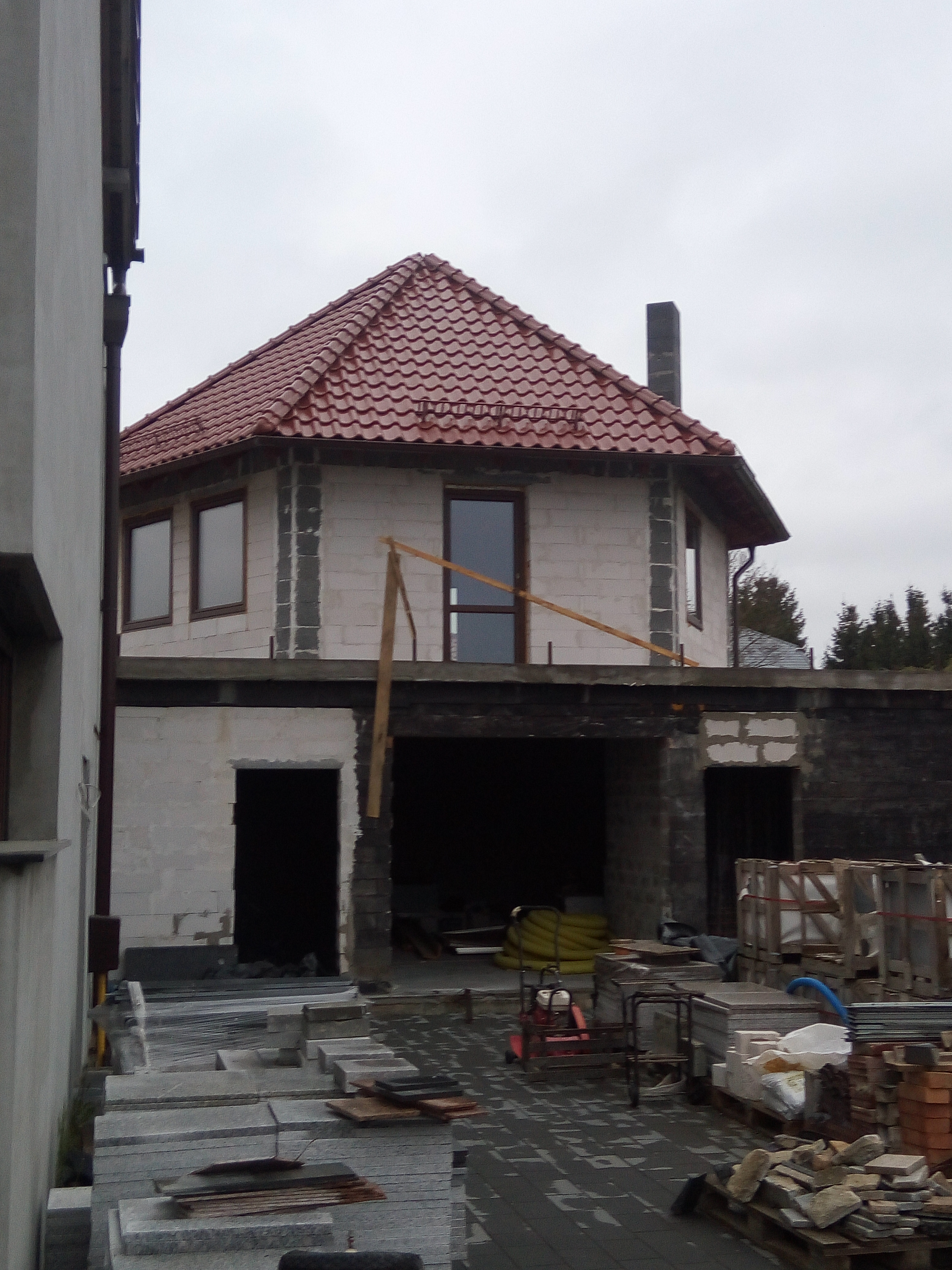 Budowa domu z czerwonym dachem z dachówki ceramicznej, widoczne pustaki, okna z drewnianą stolarką i materiały budowlane na posesji