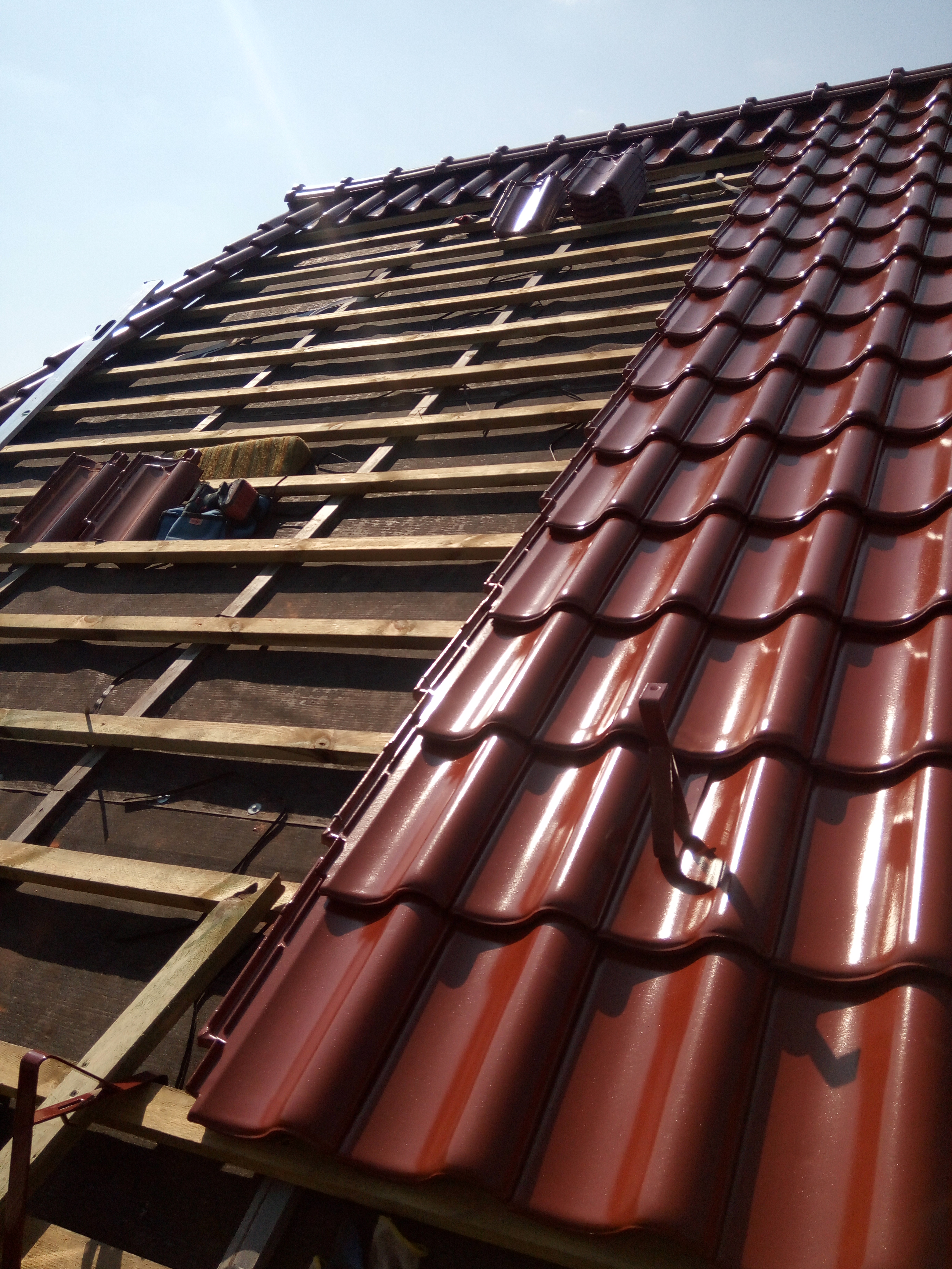 Częściowo pokryty dach z brązowej dachówki ceramicznej, widoczna drewniana konstrukcja więźby z fragmentami izolacji i narzędziami.