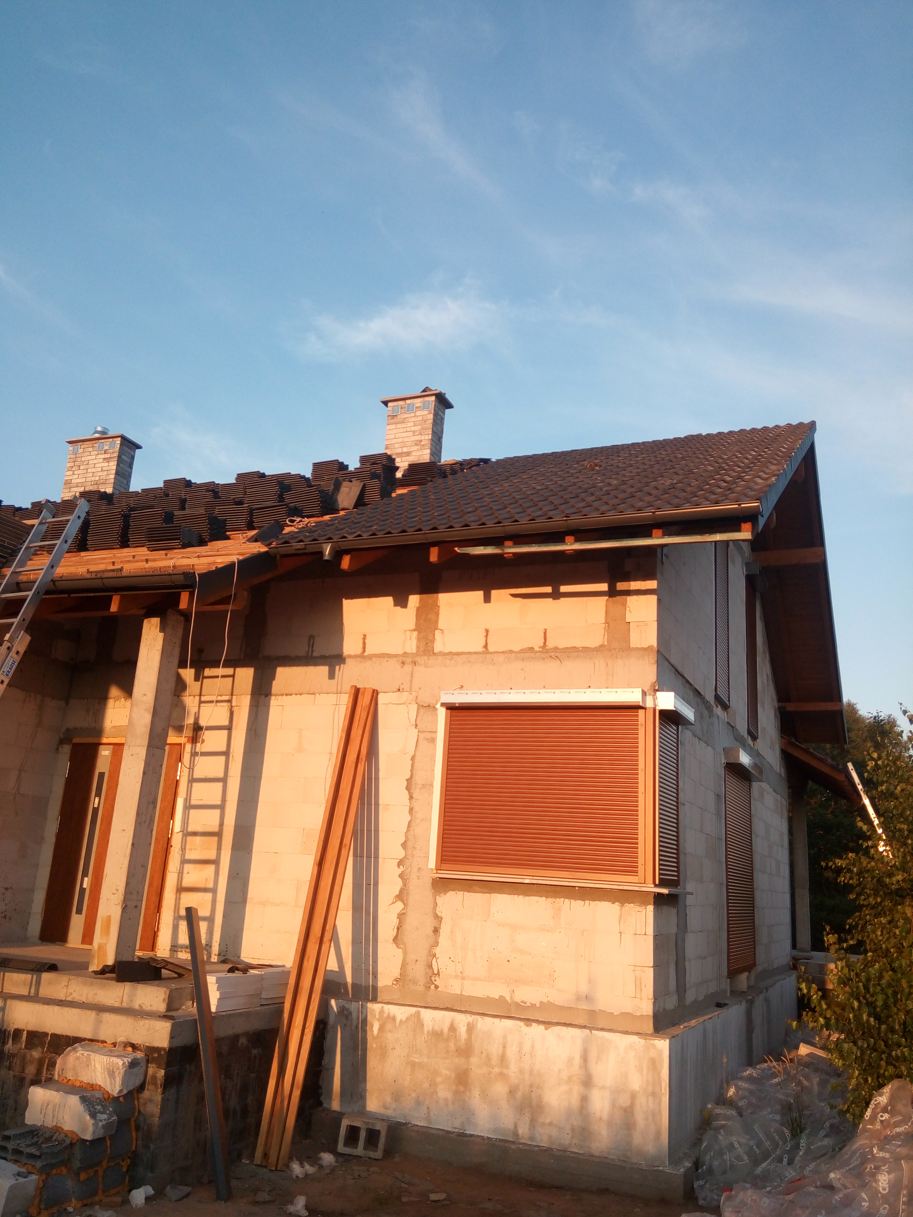 Dom w trakcie wymiany dachu z widocznymi dachówkami ułożonymi na połowie dachu, ceglanym kominem, drewnianą więźbą i rozstawioną drabiną.