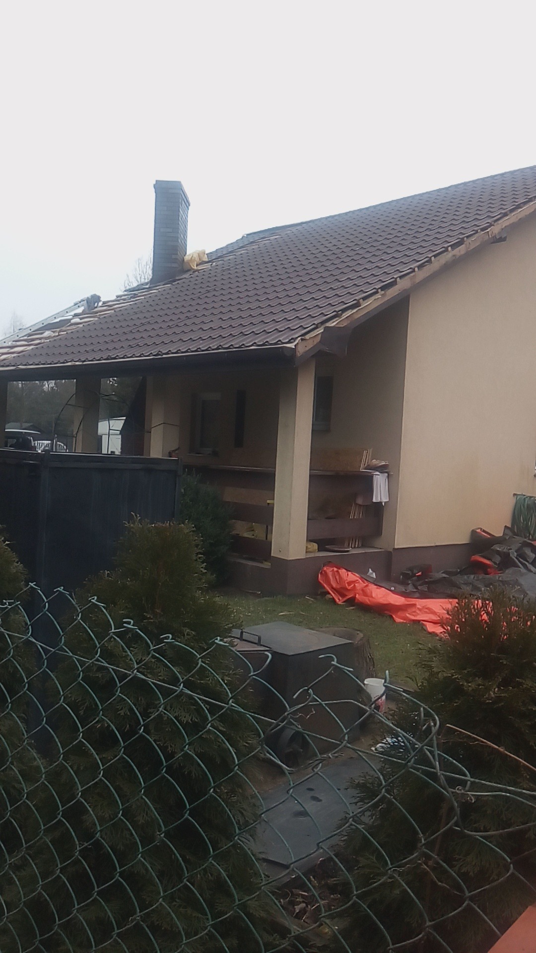 Dom jednorodzinny z częściowo zdjętym pokryciem dachowym, widoczny komin i fragment dachu z nową dachówką, zabezpieczony folią, z perspektywy ogrodu przez siatkę ogrodzeniową.
