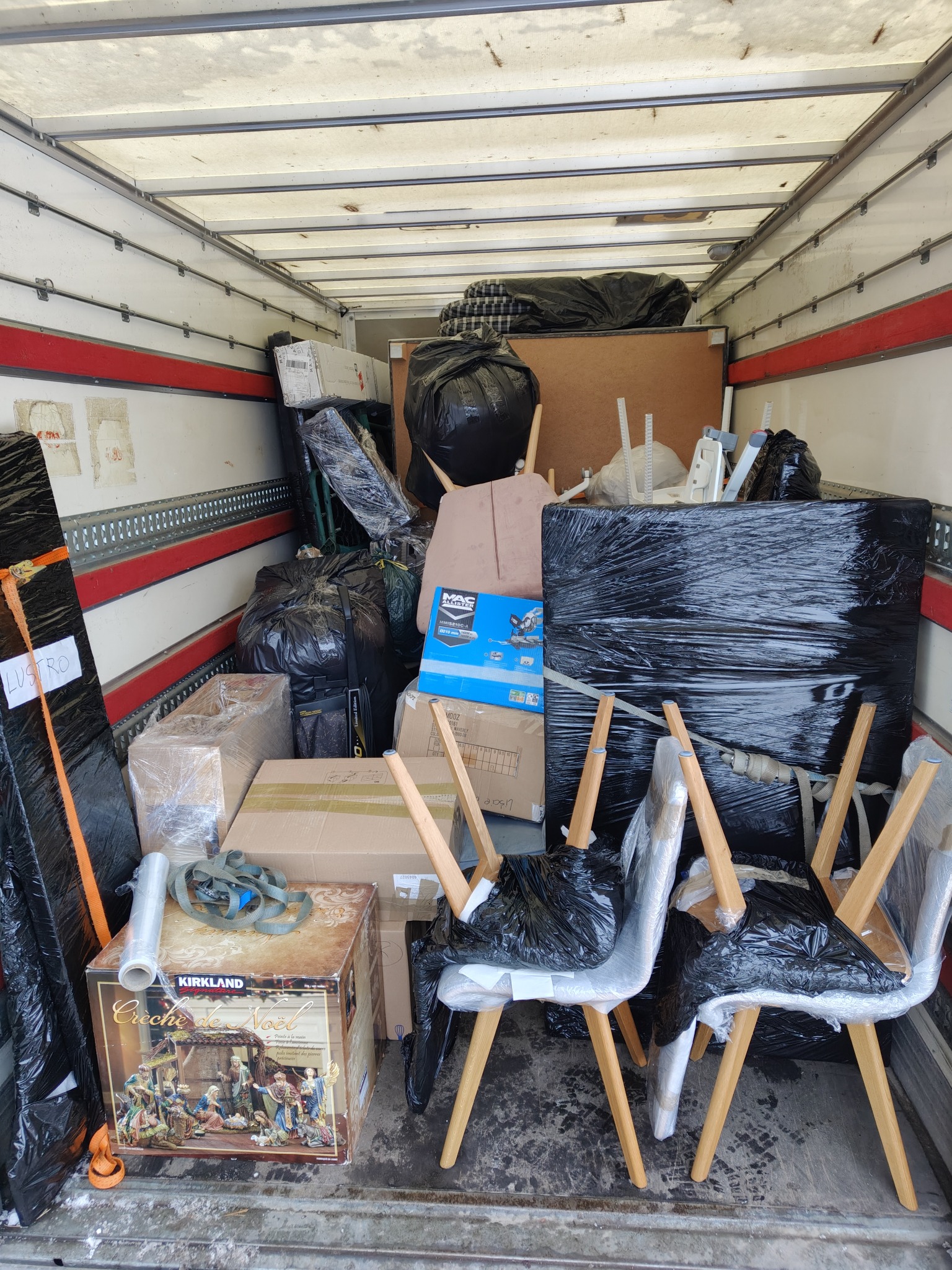 Wnętrze busa transportowego wypełnione meblami, kartonami i workami, zabezpieczonymi folią i pasami. Widoczna bożonarodzeniowa szopka Kirkland. Przeprowadzka w toku.
