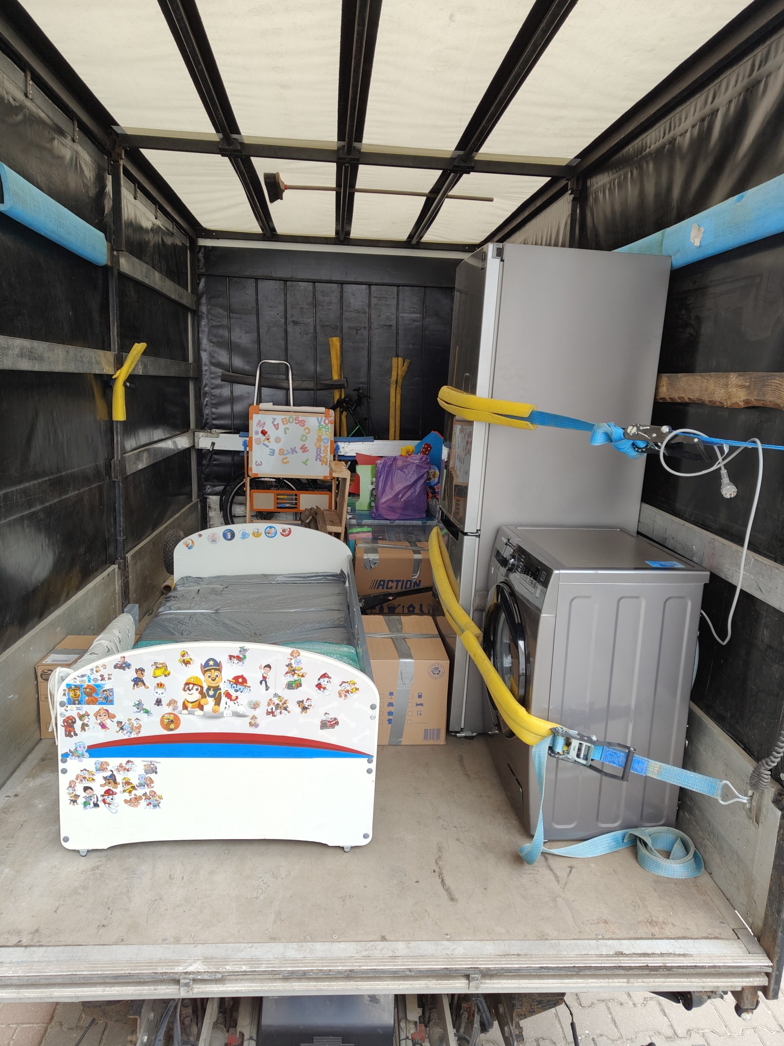 Meble i sprzęt AGD zabezpieczone pasami w przestrzeni ładunkowej busa. Widoczne łóżko dziecięce z motywem bajkowym, pralka i lodówka. Przeprowadzka.