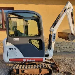 Kompaktowa minikoparka Bobcat 323, widok z przodu, zaparkowana na żwirowym podłożu przed budynkiem z oknem, ramię robocze uniesione, łyżka lekko zabrudzona ziemią.