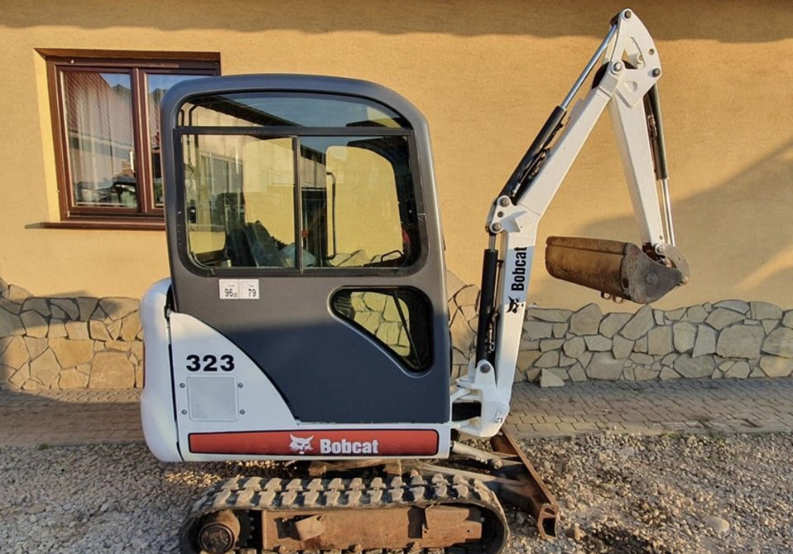 Kompaktowa minikoparka Bobcat 323, widok z przodu, zaparkowana na żwirowym podłożu przed budynkiem z oknem, ramię robocze uniesione, łyżka lekko zabrudzona ziemią.