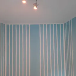 Ściana w pokoju pomalowana w jasnoniebieskie pionowe pasy oddzielone białymi paskami, z widocznym oświetleniem sufitowym.