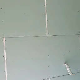 Montaż zielonych płyt kartonowo-gipsowych na ścianie, widoczne kable wystające z otworu, łączenia płyt uszczelnione masą szpachlową.