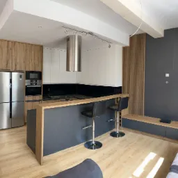 Nowoczesna kuchnia z wyspą: drewniane i białe szafki, czarny blat, stalowy okap cylindryczny, dwie czarne skórzane hokery, drewniana podłoga, szara ściana z drewnianymi panelami.