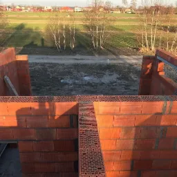 Wznoszenie ścian parteru domu z czerwonej ceramiki poryzowanej, widok z góry na budowę z polami w tle.