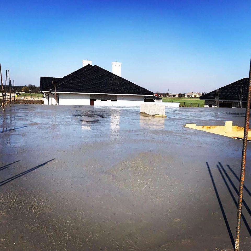 Świeżo wylana, mokra wylewka na budowie domu jednorodzinnego z widocznymi prętami zbrojeniowymi i elementami szalunku w tle, błękitne niebo.